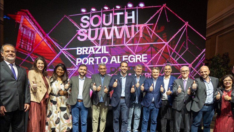 O governador no centro da imagem, rodeado à esquerda e à direita de pessoas envolvidas com o South Summit Brazil 2024. A maioria deles faz sinal de positivo com o dedo polegar e sorri. Atrás há um grande painel onde se lê: South Summit Brazil Porto Alegre.