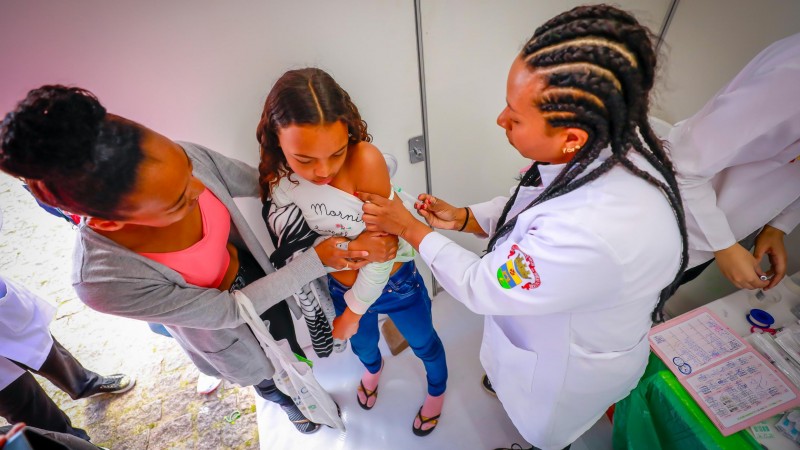 Imagem de uma t&eacute;cnica de enfermagem negra vacinando uma adolescente que &eacute; abra&ccedil;ada pela respons&aacute;vel, ambas tamb&eacute;m negras.