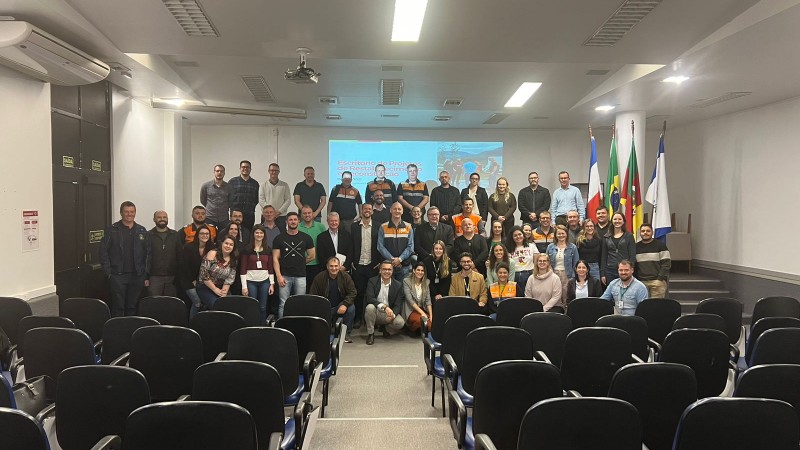 Foto posada num auditório, tirada a partir do fundo da sala. No primeiro plano, aparecem as cadeiras da plateia, de cor preta, nos cantos esquerdo e direito. Ao fundo, estão as pessoas que participaram do encontro. Elas estão distribuídas no palco, de pé. 