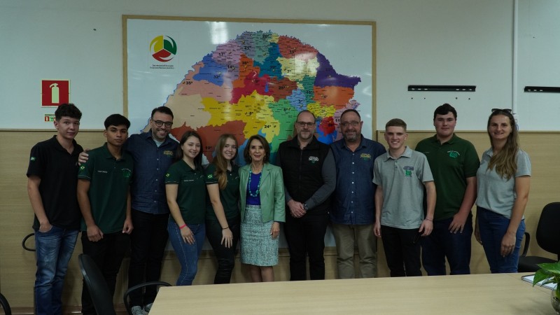 Foto em uma sala com um mapa do RS na parede ao fundo. A secretário, ao meio, posa junto aos estudantes para a câmera.