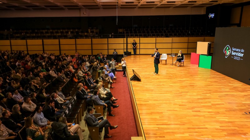 Foto do alto em que aparece à esquerda boa parte da plateia presente à abertura da Semana do Servidor 2023. Mais ao centro, de pé e ao microfone falando à plateia, está o governador Eduardo Leite. Mais à direita, está a titular da SPGG e uma enorme tela onde lê Semana do Servidor 2023.