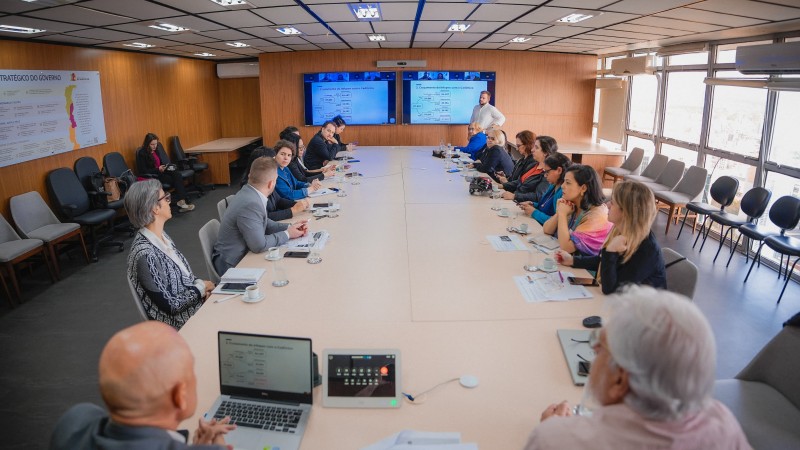 Plano geral de uma sala de reuniões, com uma longa mesa retangular de cor clara no centro, junto à qual estão sentadas várias pessoas. Ao fundo, há um homem de pé apresentando o material em duas telas acopladas a uma parede de cor marrom. À direita, há grandes janelas. 