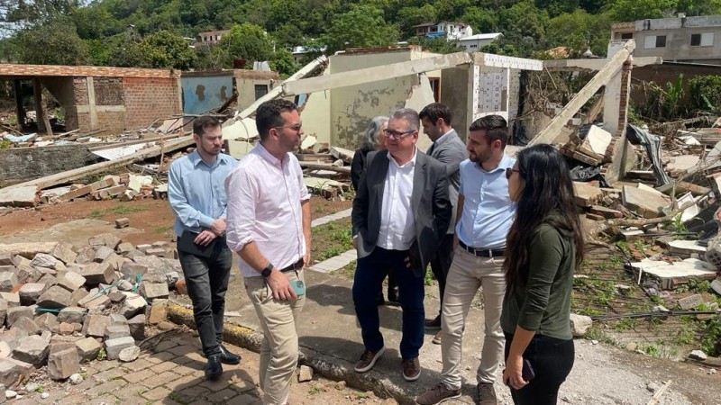 Quatro pessoas em pé formando um semicírculo e conversando. Atrás delas há mais três pessoas que não participam do diálogo mostrado na imagem. O grupo está em um ambiente aberto em uma das cidades atingidas pelas enchentes. Por isso, há escombros de casas destruídas pela força das água em todo o local.