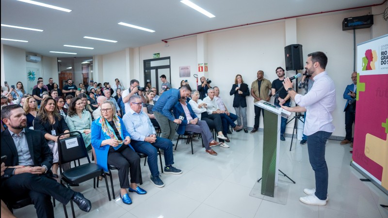 O governador Eduardo Leite discursa em um púlpito com microfone diante de uma plateia sentada em filas de cadeiras em um espaço amplo do Hospital São Camilo, em Esteio. Atrás do governador, um banner com a marca do governo.