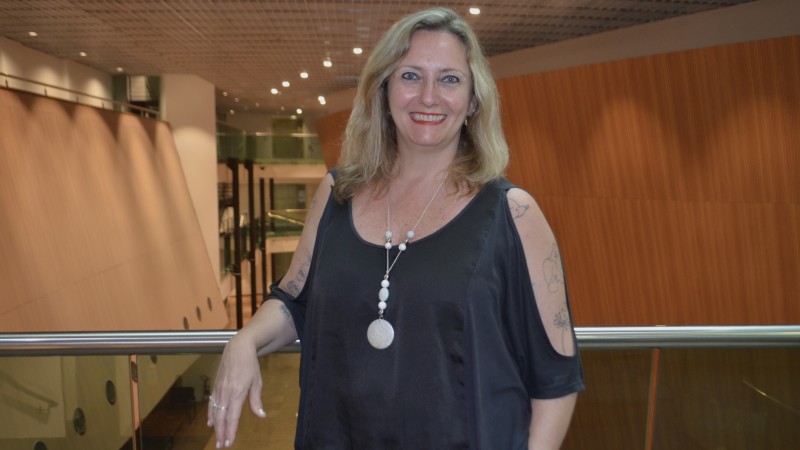 Foto posada, em plano americano. Ana está no interior de um ambiente requintado, com estruturas de madeira e luminosidade ambiente. Ela está de pé, com um cotovelo apoiado num parapeito logo atrás. Ela usa blusa preta e colar, e sorri para a câmera.