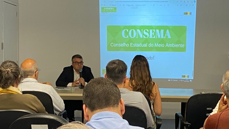 Foto em ambiente interno. Em frente a uma imagem projetada na parede, está um homem falando ao microfone, sentado em uma mesa. Na plateia, de costas, há diversas pessoas. No telão, a imagem projetada apresenta a seguinte inscrição: Consema (Conselho Estadual do Meio Ambiente).