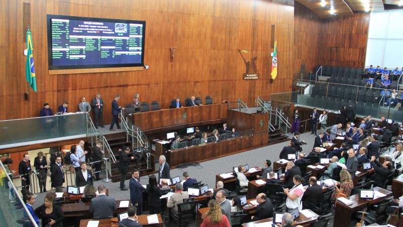 Imagem interna da Assembleia Legislativa. Diversos deputados estão na sessão, sentados ou em pé. Na frente, há um telão na parede com textos. Não é possível ler as informações.