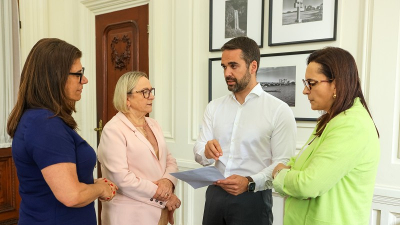 Leite em pé, falando e gesticulando em conversa com a secretária da Saúde, que está à sua esquerda, e outras duas mulheres. Leite segura em uma das mãos o documento que acabou de receber com a demanda por recursos.