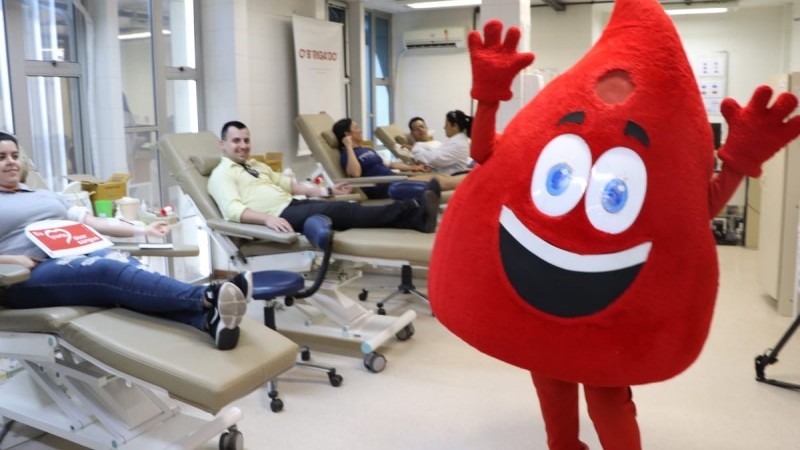 Numa sala de doação de sangue, de piso e paredes brancas, uma pessoa traja a fantasia do mascote, que consiste num corpo que emula uma grande gota de sangue. A gota tem uma cara sorridente, e a pessoa está com os braços para cima. Ao fundo, à esquerda, quatro pessoas estão deitadas em cadeiras reclináveis doando sangue.