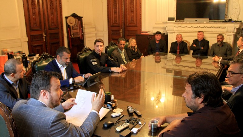 Foto em ambiente fechado. Em uma sala de reuniões, ao redor de uma mesa retangular, estão reunidas diversas autoridades em trajes formais. No canto inferior esquerdo da imagem, está o secretário-chefe da Casa Civil, com um documento em mãos, falando com os demais. 