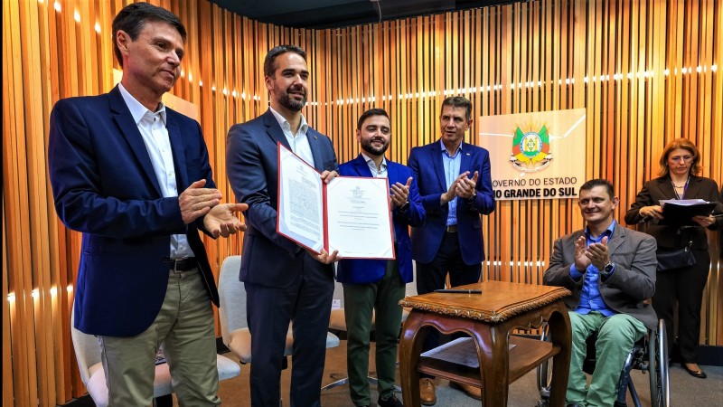 Foto posada em pé. Eduardo Leite segura e exibe o documento assinado. Ao lado dele há um homem (à esquerda) e três homens (à direita) sendo um deles cadeirante. Ao fundo, mais à direita, há uma mulher segurando uma pasta e papéis na mão.