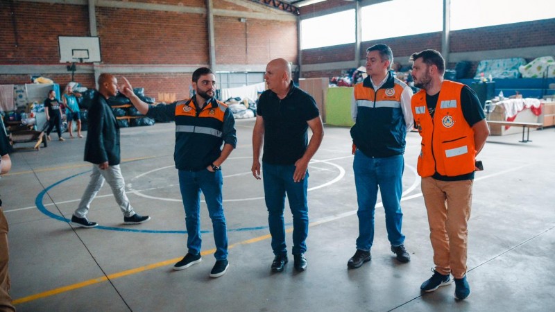 Numa quadra esportiva de ginásio, Gabriel está ao lado de mais três homens, sendo um deles o prefeito Amilton. Gabriel aponta algo com a mão, enquanto olha de lado para o prefeito.