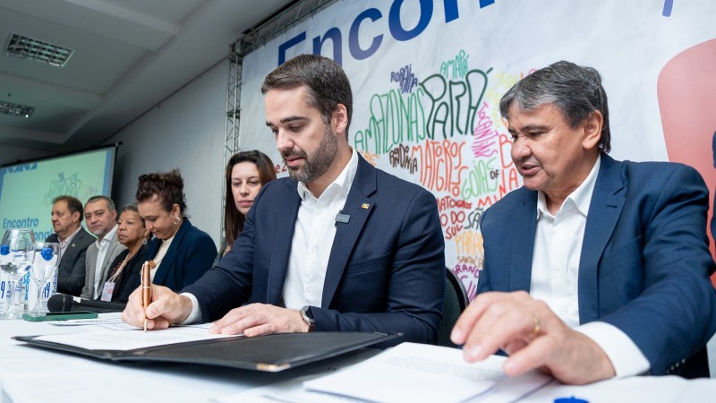 Leite assinado o documento. Ele está sentado em uma mesa formada para um evento. Do seu lado esquerdo, há três mulheres e dois homens. Do seu lado direito está o ministro Dias.