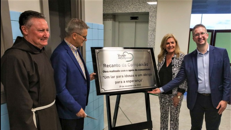 Dois homens apontam para uma placa que marca a inauguração do  Recanto da Compaixão Frei Salvador em Caxias do Sul. A placa é relativamente grande e está colocada em um cavalete. Na imagem estão presentes o frei franciscano (vestido com túnica marrom) que irá cuidar do lar de idosos e uma mulher. 