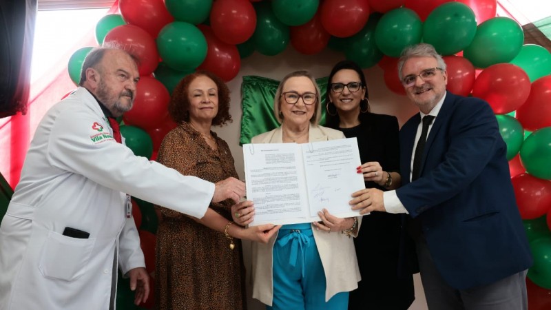 Foto posada em ambiente fechado, plano americano, de cinco pessoas. Arita está ao centro exibindo o documento. À esquerda dela, há um homem e uma mulher; à direita, também. Todos sorriem. Ao fundo, ha um conjunto de balões vermelhos e verdes em arco.