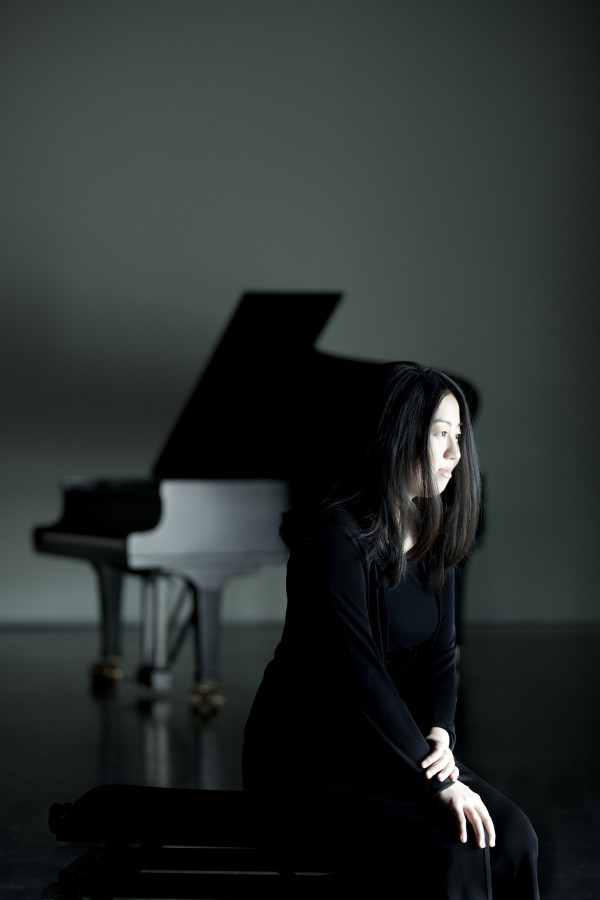 Foto artística em ambiente fechado. A pianista está sentada de lado, vestida de vestido preto. Ao fundo, há um piano preto. As paredes e o chão são escuros.