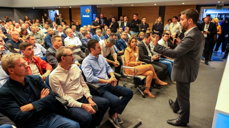 Num auditório, Leite está de pé, à direita, falando ao microfone numa mão e gesticulando com a outra. Ele está enquadrado de lado, um pouco de costas. À esquerda, aparece o público sentado. O auditório está cheio.