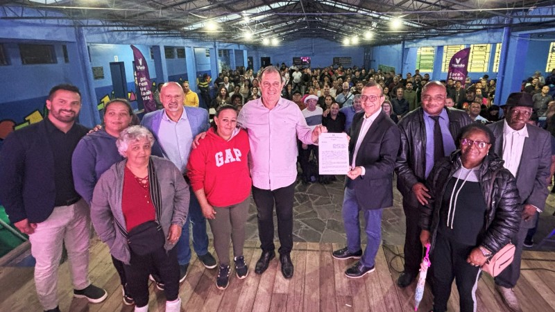 Foto posada. Dez pessoas em p&eacute; sobre um tablado e de costas para uma plateia grande. No centro o prefeito de Viam&atilde;o e o secret&aacute;rio de Habita&ccedil;&atilde;o e Regulariza&ccedil;&atilde;o Fundi&aacute;ria seguram o documento de conv&ecirc;nio que tinham acabado de assinar.