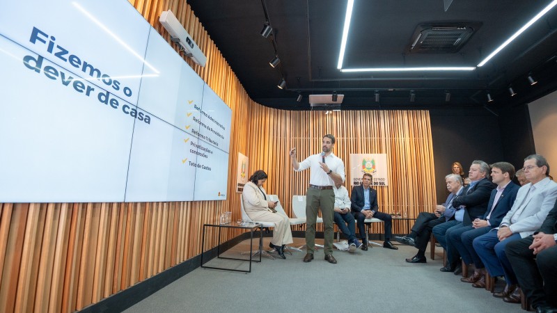 Numa sala fechada, com enquadramento levemente de cima para baixo, Leite aparece falando para uma plateia sentada em cadeiras. O ambiente é pequeno. Leite aparece de frente para a câmera, e de lado para a plateia à direita. Ele olha para a esquerda, onde está o telão com a apresentação, na qual está escrito Fizemos o dever de casa, com alguns itens.