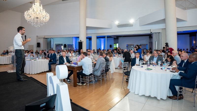 Fotografia do ambiente, que é um grande salão onde estão dispostas mesas com toalhas brancas, nas quais as pessoas estão almoçando. Leite aparece na parte esquerda da imagem, de lado para a câmera e de corpo inteiro. Ele está falando para as pessoas que almoçam e assistem. Ele está de pé e fala ao microfone. O salão tem paredes brancas e um grande lustre.