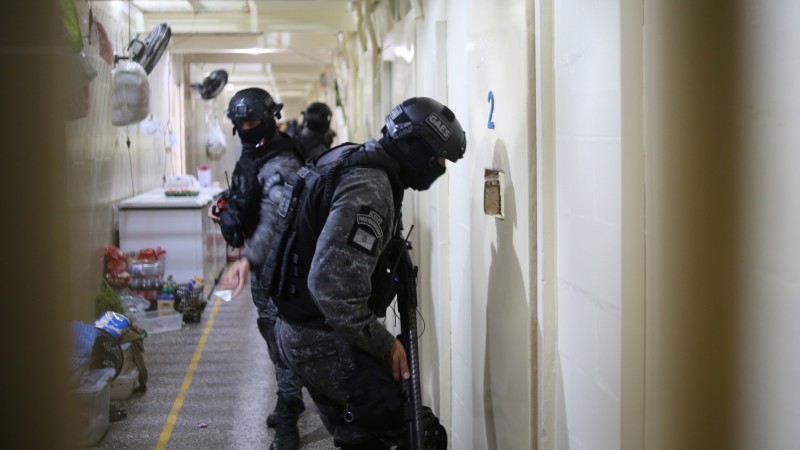 Foto tirada no interior de um pres&iacute;dio. A imagem enquadra um policial penal uniformizado com roupas escuras, com o rosto coberto, usando capacete e portando um rifle, olhando pelo v&atilde;o da porta de uma cela, de cor creme. Ele est&aacute; de lado para a c&acirc;mera. Atr&aacute;s dele, h&aacute; outros policiais.   