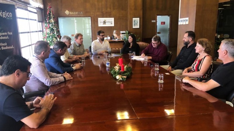 Foto numa sala de reuni&otilde;es com paredes que simulam madeira e uma grande mesa de madeira. H&aacute; v&aacute;rias pessoas sentadas ao redor da mesa, que parecem conversar. 