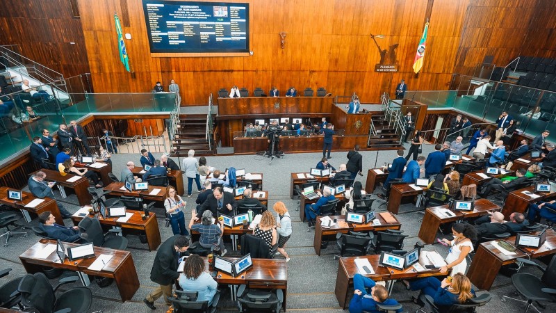 Foto do alto do Plenário da Assembleia Legislativa. Ao fundo o painel de votação e a mesa diretora e de costas na imagem os deputados em diversas mesas espalhadas pelo espaço.