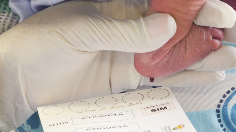 Foto em primeiro plano de uma mão com luva cirúrgica segurando o pé de um bebê. Uma gota de sangue está sando da sola do pé para cair numa cartela que serve para realizar o teste.