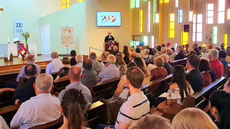 Foto em uma igreja, tirada mais do fundo, à esquerda. O público aparece de costas, sentado, assistindo ao ato. Uma liderança conduz a fala no púlpito.