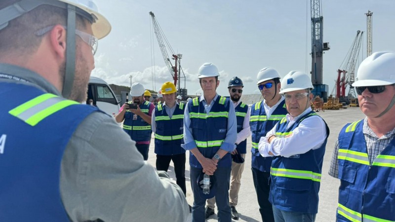 Foto ao ar livre, em alguma área do complexo portuário. A comitiva usa equipamentos de proteção (capacete branco e colete azul) e acompanha a fala de um técnico. Aparecem oito pessoas na foto, de pé.