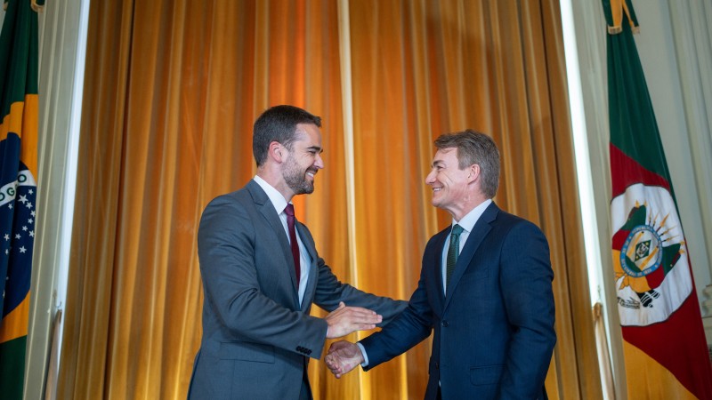 Foto em ambiente fechado, com foco no governador Eduardo Leite e no deputado Vilmar Zanchin, que transmitiu o cargo. Eles estão sorrindo e se cumprimentando, de frente um para o outro. Ao fundo, há uma cortina dourada e as bandeiras do Brasil à esquerda e do Rio Grande do Sul à direita. 