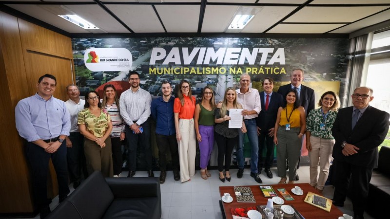 Foto posada em ambiente fechado. 17 pessoas estão de pé olhando para a câmera, a maioria sorrindo. Ao centro os secretários seguram o documento assinado