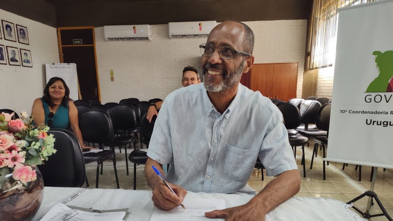 Foto em close de um homem negro, calvo e de óculos que está sentado diante de uma mesa com uma caneta em uma das mãos assinando papéis. Atrás dele, estão sentadas duas pessoas que sorriem. Ele também sorri.