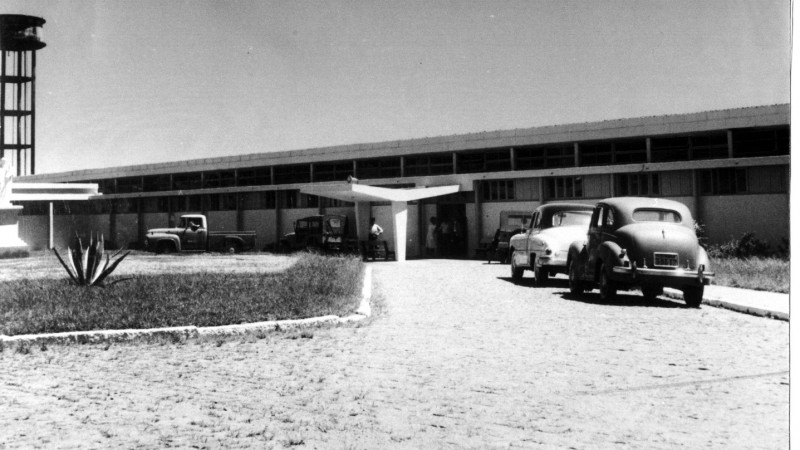 Foto antiga, em preto e branco, da fachada do hospital, que é um estabelecimento verticalizado. A imagem enquadra ainda o caminho dos carros para parar em frente a ele. Há dois carros estacionados. 