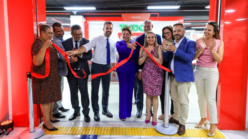 Foto em ambiente fechado, com em torno de dez pessoas realizando o descerramento da faixa de inauguração, de cor vermelha. No centro, está o governador, que solta a faixa enquanto sorri.