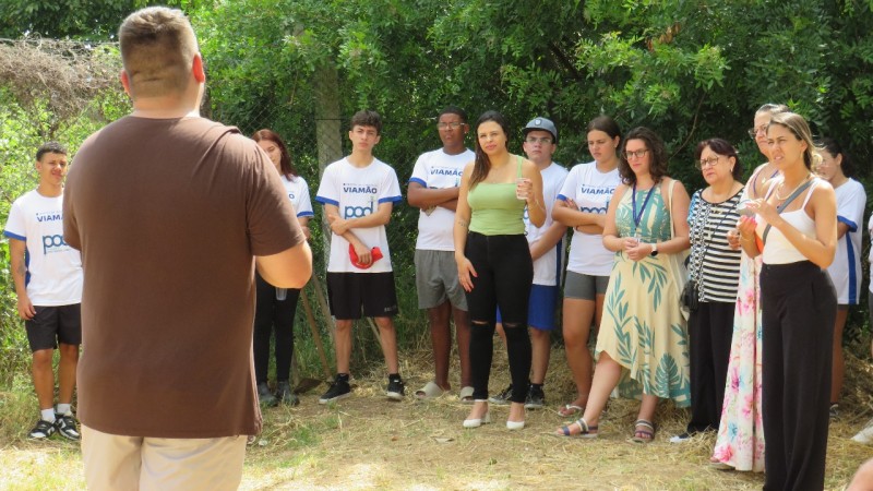 Foto em ambiente aberto de pessoas enfileiradas, de pé, acompanhando um homem que está de costas para a câmera e fala aos presentes. O local é de natureza, com presença de árvores ao fundo.