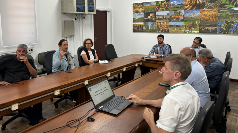 Foto em sala de reuniões de paredes brancas. Oito pessoas estão sentadas em uma mesa retangular, enquanto conversam.