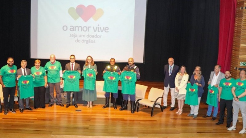 Foto posada. Diversas pessoas em pé em um palco segurando a camiseta da campanha O Amor Vive, que também aparece projetada atrás das pessoas em um grande painel.