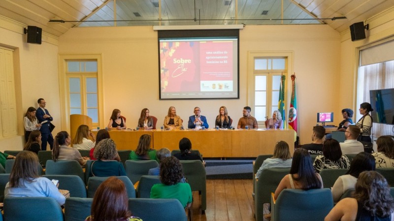 Foto da plateia de costas diante de um tablado com uma enorme mesa, atrás da qual estão sentados dois homens e cinco mulheres compondo a mesa do evento. Atrás deles, na parede, há um painel na parede, sobre o qual está projetado algo euq não pode ser lido.