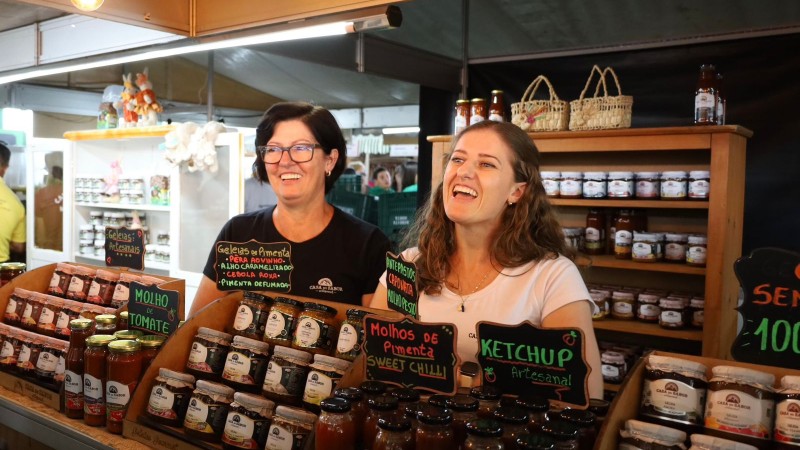 Duas mulheres que lideram empreendimento da agricultura familiar sorrindo atrás de um balcão cheio de produtos em um estande do Pavilhão da Agricultura Familiar na Expodireto Cotrijal 2024