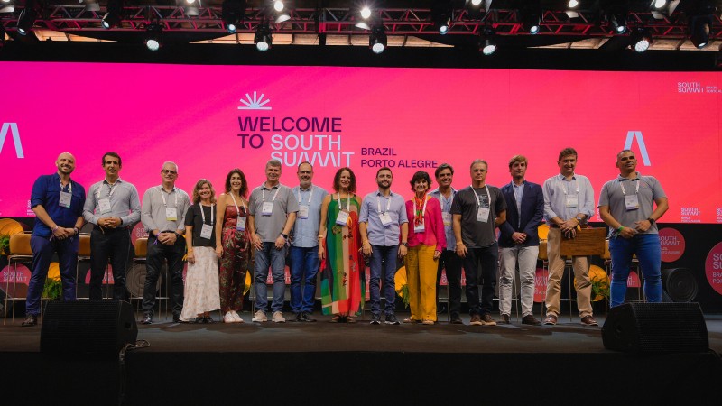 Foto posada. Onze homens e quatro mulheres em p&eacute; sobre um palco. Ao fundo est&aacute; um painel no qual est&aacute; escrito: Welcome to South Summit Brazil.