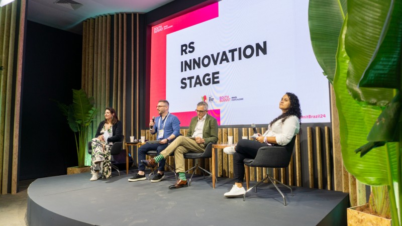 Duas mulheres e dois homens sentados em cadeiras sobre um tablado. Um deles fala ao microfone. Atrás deles uma tela exibe as palavras RS Innovation Stage.