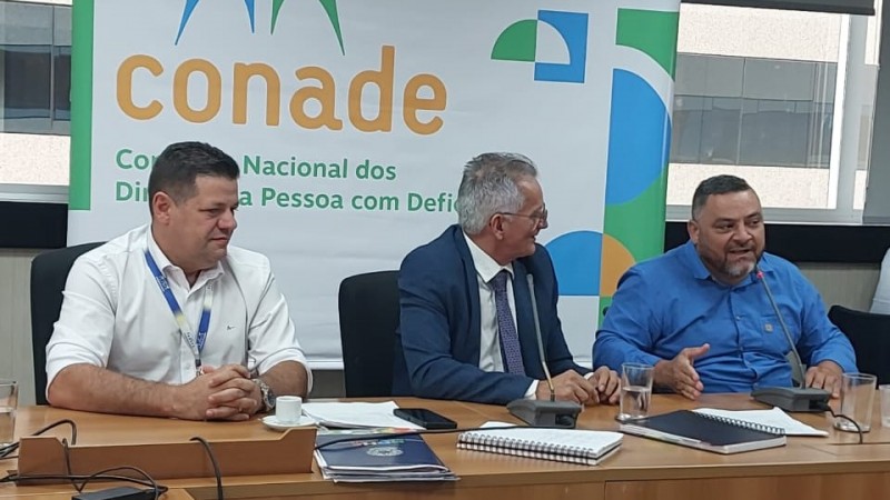 Foto de três homens sentados junto a uma mesa de madeira. À direita, Marquinho fala ao microfone. Ao fundo, um painel escrito Conade.