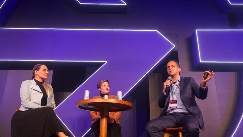 Foto dos três participantes sentados na apresentação; um deles fala ao microfone. O fundo é escuro com um símbolo de cor roxa.