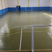 Foto geral de uma quadra poliesportiva. O espaço é fechado, o piso é verde-musgo, e há traves de gol de futebol instaladas.