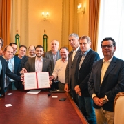 Gabriel segura o documento recém assinado para o setor Leiteiro rodeado por 12 homens. A foto é posada em um ambiente dentro do Palácio Piratini onde ocorreu a reunião.
