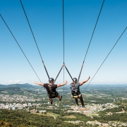 Imagem de duas pessoas de costas descendo de tirolesa em Feliz. Na imagem, a cidade pode ser vista do alto e à distância.