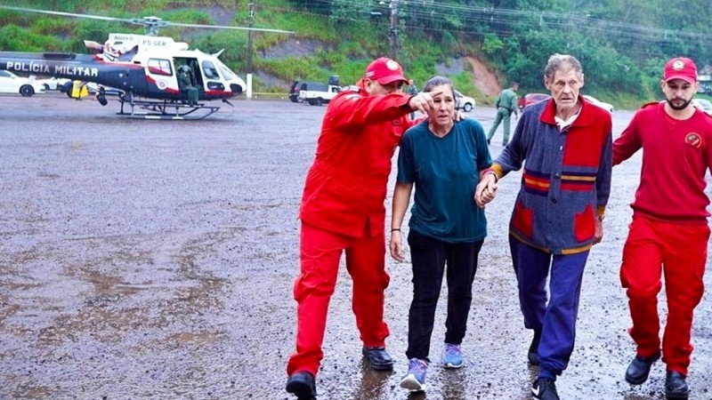 Casal acabando de ser resgatado em São Sebastião do Caí> eles caminha acompanhados por dois bombeiros. Ao fundo, pode ser visto o helicóptero.