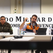 Leite, o prefeito de Porto Alegre e o coronel Hertz sentados atrás de uma mesa na coletiva sobre enchentes na sexta 3 de maio. Leite, com o microfone na mão, fala e gesticula. Atrás deles há um painel onde se lê Comando Militar do Sul.