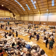 Cete recebendo desalojados de Porto Alegre devido a enchentes. Imagem aberta e do alto do interior do ginásio, no qual há diversas pessoas, além de objetos e colchões espalhados pelo chão.